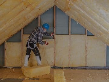 Contractor installing fiberglass insulation between wall studs inside an unfinished attic space with exposed wooden beams and framing.