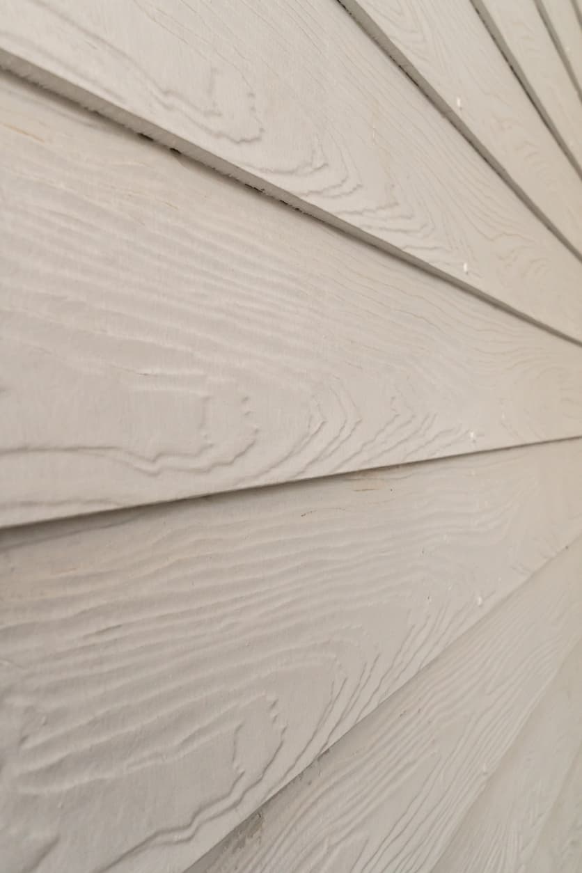 Close-up angled view of light beige fiber cement house siding panels with visible wood grain texture and overlapping horizontal boards.