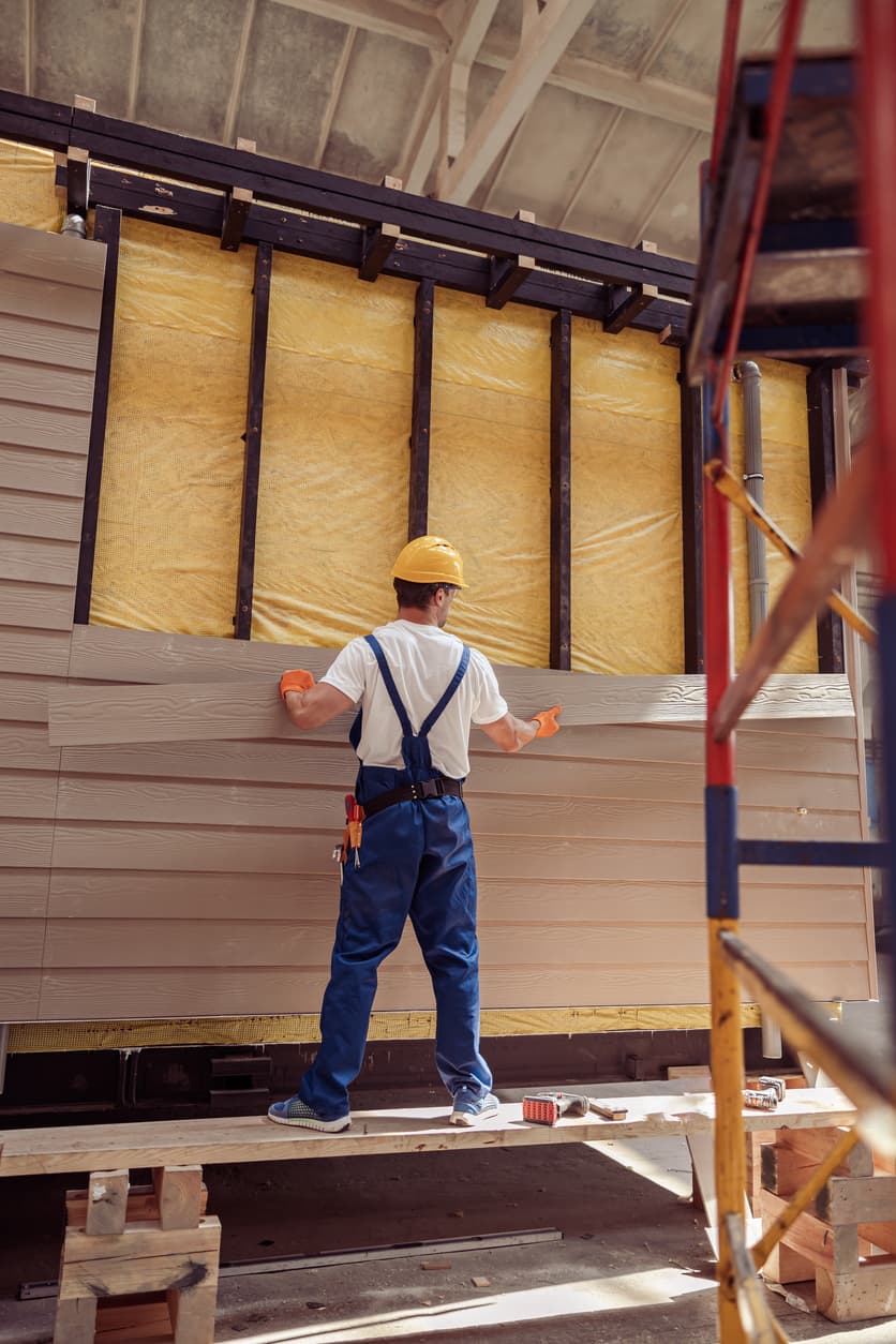 Construction worker installing horizontal exterior siding panels on a building wall with exposed insulation and framing at a residential construction site.