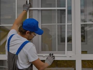 Contractor wearing safety gloves and a blue cap installing a white-framed vinyl window on a residential home exterior.