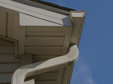 Close-up view of a residential roof edge showing white gutters, downspout, soffit vents, and fascia trim against a clear blue sky.