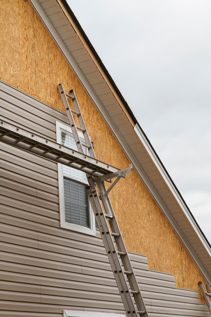 Extension ladders positioned against a two-story house during exterior siding installation, with exposed wall sheathing and partially completed vinyl siding beneath roof soffits.