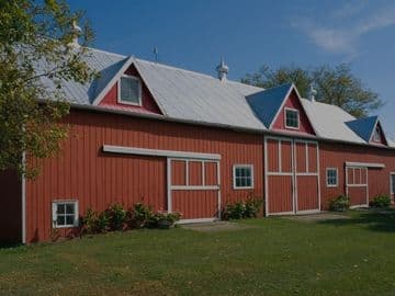 Large red barn with white trim and board-and-batten siding, featuring multiple doors and a metal roof on a grassy rural property under a blue sky.