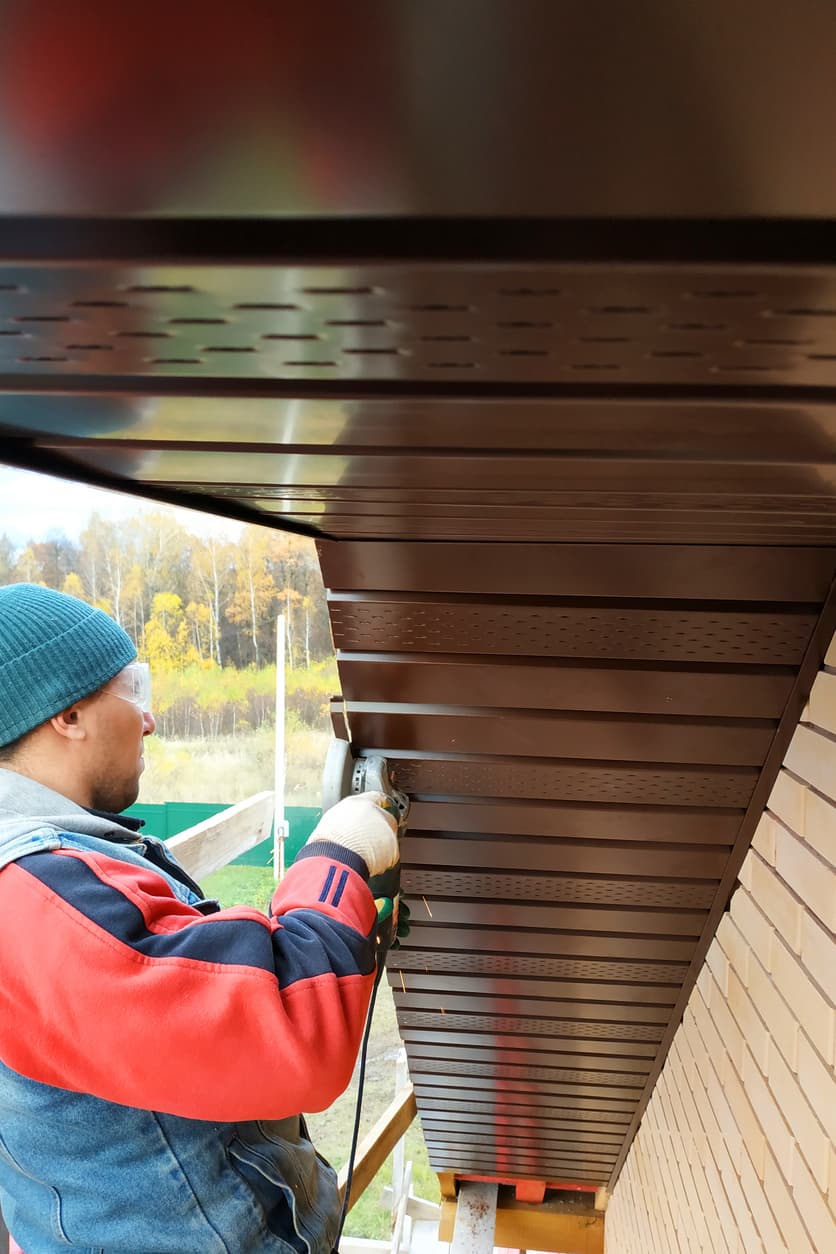 soffit-installation-home-exterior-work Construction worker installing or repairing metal soffit panels beneath a residential roof overhang using a power tool.